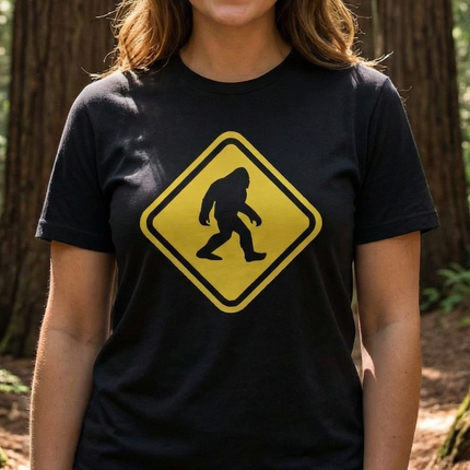 Unisex black t-shirt featuring a yellow Sasquatch crossing warning sign graphic.