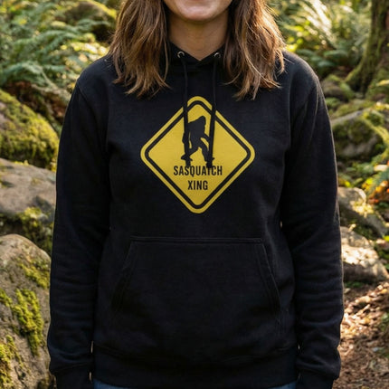 Yellow Sasquatch crossing warning sign graphic on a black pullover hoodie.